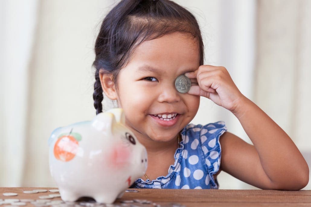 A little girl in front of her piggy bank smiling while holding up a quarter to her squinted eye